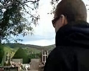 Uma pessoa vestindo um moletom escuro e óculos fica ao ar livre, de frente para uma paisagem com árvores e colinas sob um céu nublado.