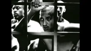 Uma imagem monocromática do Mano Brown e outros homens atrás das grades, segurando o metal com as mãos.