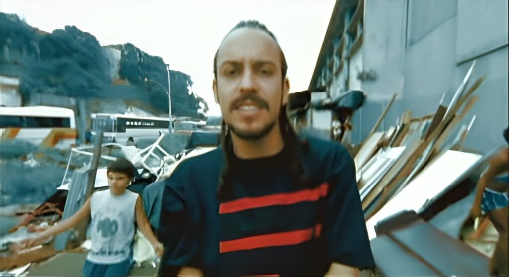 Gabriel o Pensador em destaque com uma camiseta preta com listras vermelhas; no fundo, pessoas buscam coisas em meio a entulho de móveis e afins.