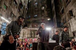 Ice Blue, Mano Brown, Edi Rock e Dexter aparecem em cima de um palco, rodeado de pessoas e construções prediais visivelmente desgastadas. Mano Brown segura o microfone. Eles estão se apresentando em uma Ocupação em São Paulo.