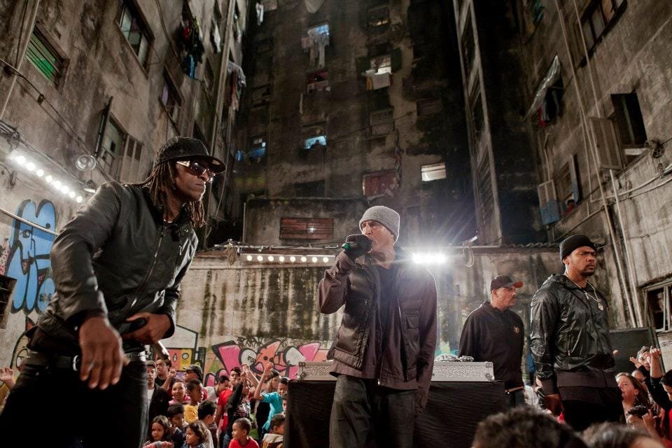 Ice Blue, Mano Brown, Edi Rock e Dexter aparecem em cima de um palco, rodeado de pessoas e construções prediais visivelmente desgastadas. Mano Brown segura o microfone. Eles estão se apresentando em uma Ocupação em São Paulo.