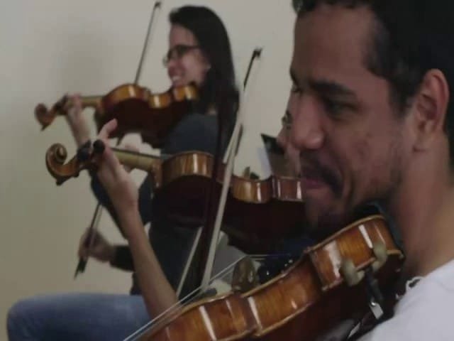 Homem em destaque e mulher ao fundo ambos tocando violino na Orquestra Sinfônica Heliópolis.