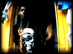 Sabotage com uma camisa preta com a sua imagem em branco igual na capa do disco Rap é Compromisso em uma porta de um barraco de madeira amarela na sua quebrada.