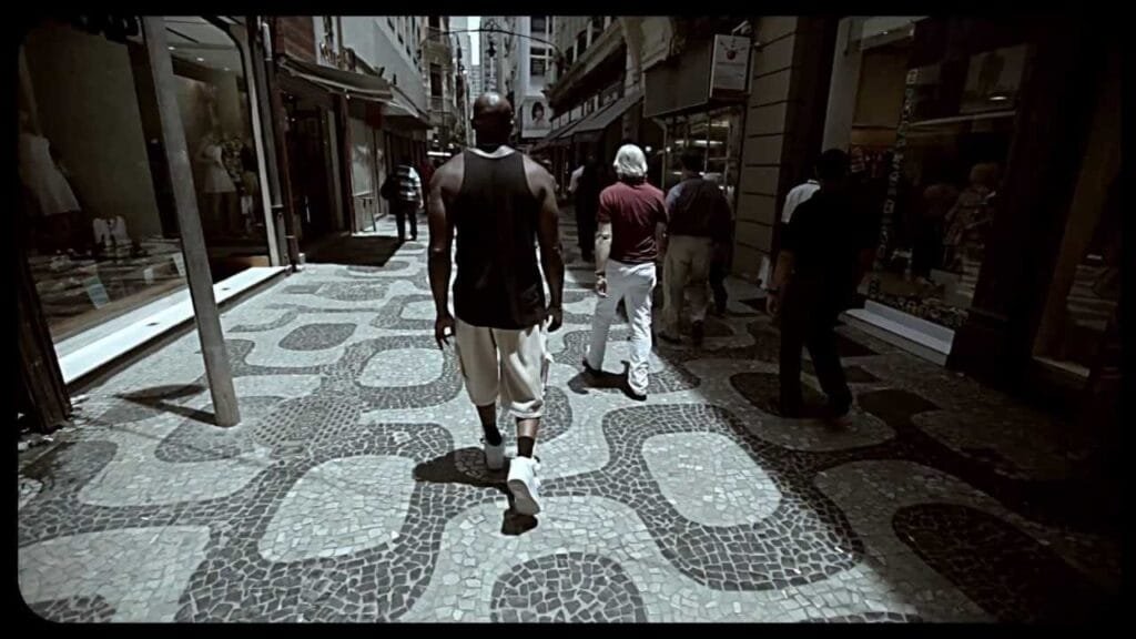 A imagem mostra a vista de trás de um homem, que parece ser MV Bill, caminhando em uma rua movimentada. Ele está vestindo uma camiseta regata preta e calças claras. A rua tem um calçamento característico de pedras portuguesas, com padrões geométricos escuros e claros. Ao longo da rua, há edifícios com lojas e vitrines de ambos os lados. Outras pessoas podem ser vistas caminhando na mesma direção ou em sentido contrário ao homem em destaque. A iluminação sugere que é dia, e a cena tem um aspecto de rua urbana, possivelmente no Brasil, dado o tipo de calçamento. A imagem é enquadrada com bordas escuras, como se fosse um trecho de um vídeo ou filme.