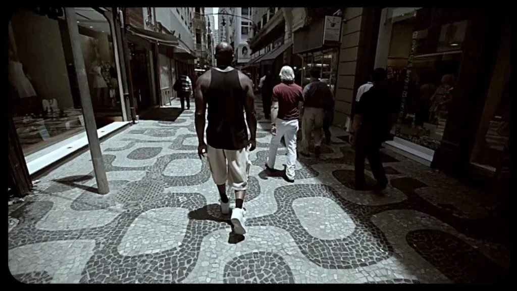 A imagem mostra a vista de trás de um homem, que parece ser MV Bill, caminhando em uma rua movimentada. Ele está vestindo uma camiseta regata preta e calças claras. A rua tem um calçamento característico de pedras portuguesas, com padrões geométricos escuros e claros. Ao longo da rua, há edifícios com lojas e vitrines de ambos os lados. Outras pessoas podem ser vistas caminhando na mesma direção ou em sentido contrário ao homem em destaque. A iluminação sugere que é dia, e a cena tem um aspecto de rua urbana, possivelmente no Brasil, dado o tipo de calçamento. A imagem é enquadrada com bordas escuras, como se fosse um trecho de um vídeo ou filme.