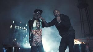 A imagem mostra MC Cidinho e MV Bill em um ambiente escuro e noturno, com fumaça ou névoa no ar e luzes desfocadas ao fundo. À esquerda, MC Cidinho usa um boné escuro, óculos escuros e uma camiseta estampada com uma foto do Bené, de Cidade de Deus. Ele tem a mão direita levantada perto do rosto. À direita, MV Bill, com barba e vestindo um moletom escuro e correntes no pescoço, está com um braço estendido, apontando para frente com o dedo indicador. Há um barril com fogo visível no lado direito da imagem.
