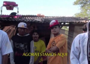 A imagem mostra um grupo de pessoas em frente a uma casa simples, possivelmente em uma área rural ou suburbana. No primeiro plano, à esquerda, há um homem usando um moletom preto com um boné branco virado para trás. Ao lado dele, um pouco mais para trás, outra pessoa veste uma camiseta amarela e tem a boca aberta, como se estivesse falando ou cantando. No centro da imagem, um homem de boné vermelho e branco e camiseta laranja está com os braços cruzados, olhando diretamente para a câmera. Há um balão de texto na parte inferior da imagem que diz 'AGORA ESTAMOS AQUI'. A casa atrás deles tem um telhado de amianto e paredes que parecem ser de madeira ou tábuas. A vegetação pode ser vista no fundo, à direita.