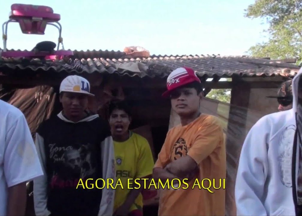 A imagem mostra um grupo de pessoas em frente a uma casa simples, possivelmente em uma área rural ou suburbana. No primeiro plano, à esquerda, há um homem usando um moletom preto com um boné branco virado para trás. Ao lado dele, um pouco mais para trás, outra pessoa veste uma camiseta amarela e tem a boca aberta, como se estivesse falando ou cantando. No centro da imagem, um homem de boné vermelho e branco e camiseta laranja está com os braços cruzados, olhando diretamente para a câmera. Há um balão de texto na parte inferior da imagem que diz 'AGORA ESTAMOS AQUI'. A casa atrás deles tem um telhado de amianto e paredes que parecem ser de madeira ou tábuas. A vegetação pode ser vista no fundo, à direita.