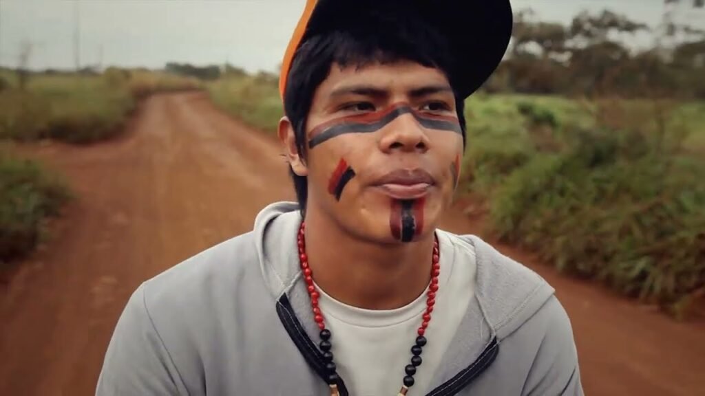 Uma imagem de close de um jovem indígena, com o rosto pintado em padrões tribais nas cores preto, vermelho e branco. Ele usa um boné virado para trás, uma blusa de moletom cinza com capuz e dois colares de contas, um vermelho e outro preto. A expressão facial do jovem é neutra, com a língua levemente para fora. Ao fundo, uma estrada de terra em tom avermelhado se estende por um campo gramado e árvores, sob um céu nublado.