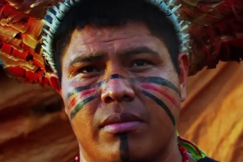 Uma imagem em close de um homem indígena com feições sérias, olhando diretamente para frente. Ele usa um cocar com penas em tons de marrom e bege. Seu rosto está pintado com traços largos em preto, vermelho e um fino traço amarelo, passando pelas bochechas e descendo pelo centro do queixo. Sua pele é morena e o fundo está desfocado em tons quentes de laranja e marrom.
