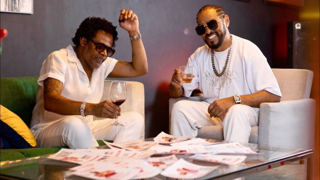 A imagem mostra dois homens sentados em poltronas confortáveis, em um ambiente que parece ser um lounge ou sala de estar. À esquerda, o cantor Xande de Pilares, um homem de cabelo crespo escuro e óculos de sol, veste uma camisa branca de manga curta e calças brancas. Ele segura um copo com vinho tinto na mão esquerda, enquanto a mão direita está levantada, com os dedos dobrados como se estivesse fazendo um gesto. No braço, ele usa uma pulseira prateada. À direita, o rapper Thaide usando óculos de sol escuros, veste uma camiseta branca larga e calças brancas. Seus cabelos estão em tranças finas. Ele segura um copo com uma bebida âmbar na mão direita e exibe um sorriso largo. Em seu pescoço, há uma corrente dourada e, no pulso esquerdo, um relógio de pulso. Entre os dois, há uma mesa de centro de vidro coberta por vários papéis e revistas espalhados. Ao fundo, percebe-se uma parede escura e, à esquerda, parte de um sofá verde com uma almofada amarela. A iluminação é suave e uniforme, criando uma atmosfera relaxada.