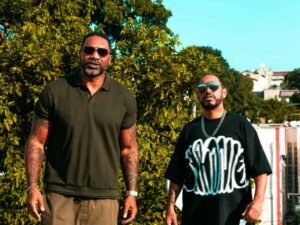 Dois homens, os rappers MV Bill (à esquerda) e Thaíde (à direita), posando para uma foto em um dia ensolarado. MV Bill veste uma camisa polo verde musgo de manga curta e óculos de sol, com braços tatuados visíveis. Thaíde usa uma camiseta preta com uma estampa branca marcante, corrente grossa no pescoço e óculos de sol. Atrás deles, há uma vasta folhagem verde e, mais ao fundo, algumas construções e o céu azul.