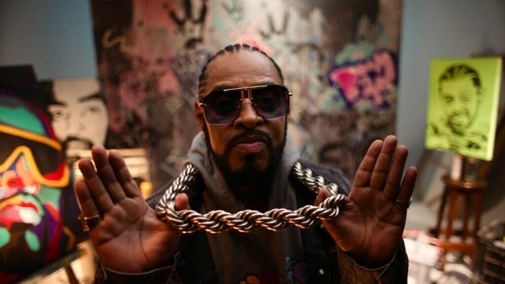 A imagem mostra o rapper brasileiro Thaíde, um homem negro com barba e tranças, usando óculos de sol escuros e uma corrente grossa e prateada em suas mãos, exibindo-a para a câmera. Ele está em um ambiente que parece ser um estúdio ou galeria de arte, com quadros e grafites abstratos ao fundo, em tons de verde, cinza e roxo; é possível distinguir, entre as artes ao fundo, pinturas de retratos do próprio rapper. A iluminação destaca o artista e a corrente.