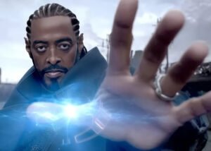 A imagem é um close-up de Thaíde, um homem de meia-idade com a pele escura, barba e cabelo com tranças curtas. Ele está usando uma roupa escura que lembra uma capa ou casaco. Seus olhos estão arregalados e fixos na câmera, com um olhar intenso. Sua mão direita está estendida em primeiro plano, com a palma voltada para a câmera, e anéis podem ser vistos em seus dedos. Há um efeito de luz azul brilhante e eletrizante emanando de sua palma, sugerindo algum tipo de poder ou energia. O fundo está desfocado e parece ser um cenário urbano ou industrial.