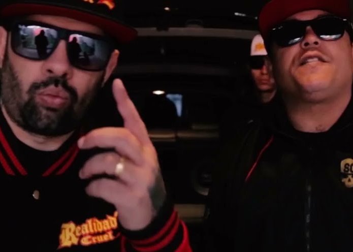 DJ Bola8 e WGI no clipe de Eterno Malokeiro. Ambos estão usando óculos escuros, bonés e jaquetas escuras. Bola8, à esquerda, tem barba, está apontando um dedo para cima e usa um anel dourado. WGI, à direita, tem bigode e está olhando para a frente. Outra pessoa, menos visível, está atrás deles.