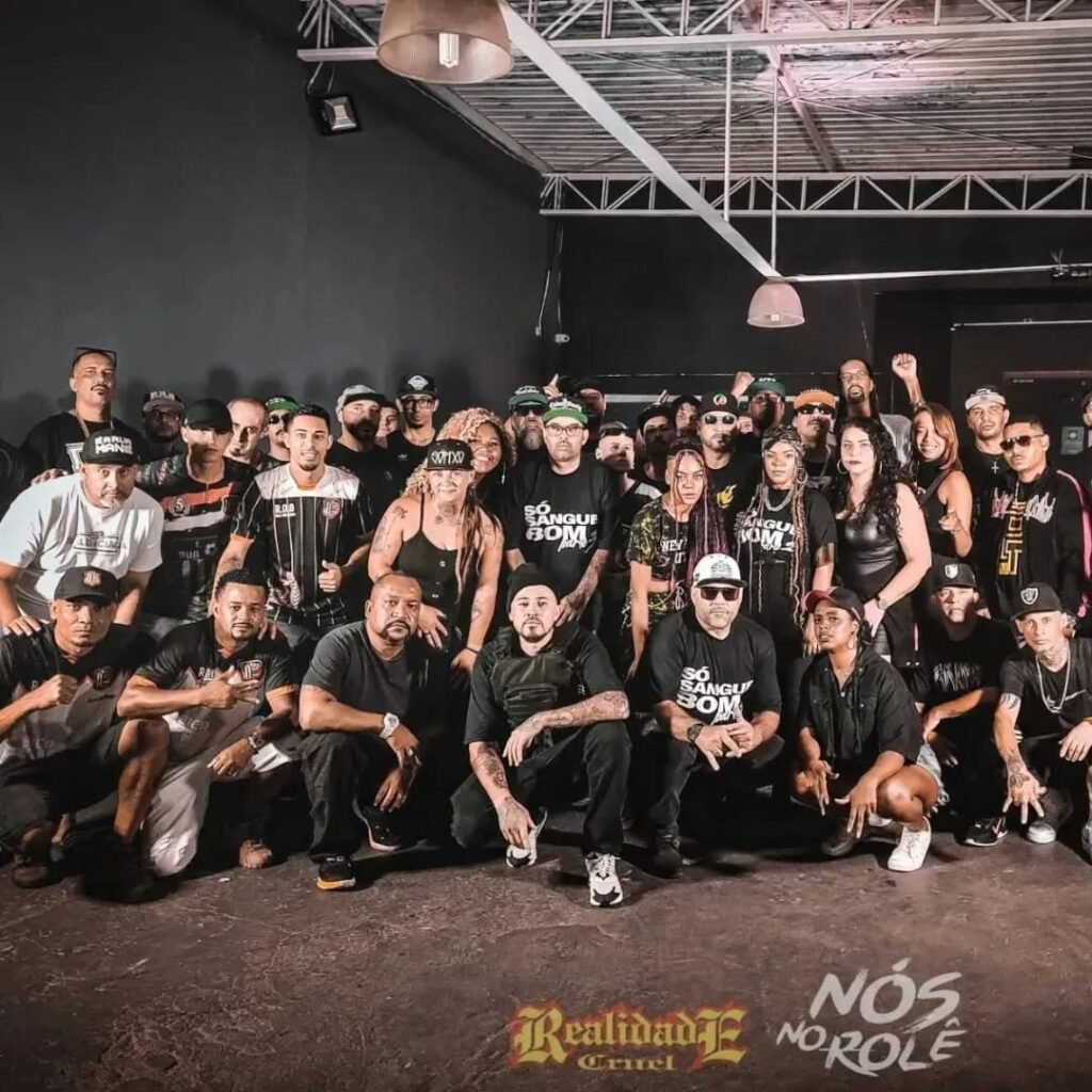 Uma grande foto de grupo, com cerca de 30 pessoas que participaram das filmagens do clipe Sangue Bom (Parte 3), em um ambiente interno escuro. Os artistas estão posando juntos, a maioria em pé e alguns agachados na frente. Todos estão usando roupas escuras e muitos com uma camisa temática com o título da música. No canto inferior da imagem, há o logo do grupo 'Realidade Cruel' e o texto 'NÓS NO ROLÊ', que foi a equipe responsável pelas fotos da gravação do clipe.