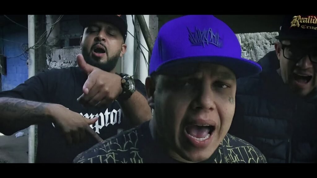 Três homens em um ambiente urbano, possivelmente industrial. Nocivo Shomon em primeiro plano, usando um boné roxo, tem uma expressão de choque ou está gritando. Léo RC atrás dele e à esquerda gesticula com o polegar para cima. TM Thiago Mascote é visível à direita, também com uma expressão de choque ou gritando. Todos os três homens usam bonés.