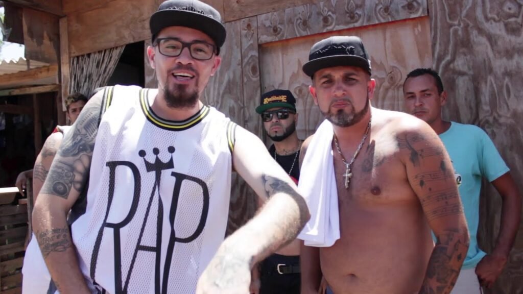 TM Thiago Mascote de óculos e boné para trás, usando uma camisa regata branca de basquete com os dizeres 'RAP', apontando para a câmera. Ao lado dele, Chicão aparece sem camisa com várias tatuagens nos braços, uma toalha branca no pescoço e um boné. Ao fundo, R.O. e outros dois homens em frente a uma parede de madeira.