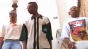 DJ Kri, Athalyba Man e Grand Master Duda em plano médio, olhando para baixo, em uma cena do clipe 'Política'. Athalyba Man, ao centro, veste uma camisa polo de manga curta verde escura e creme. Grand Master Duda, à direita, veste uma camiseta branca estampada e óculos. DJ Kri, à esquerda, veste uma polo creme e jeans. A cena externa tem uma parede clara e detalhes arquitetônicos.