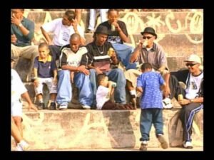 Foto do clipe 'Tik Tak', do Doctor Mc's, com os integrantes do grupo em meio jovens e alguns meninos sentados em uma arquibancada de cimento pichada, sob a luz do sol. As pessoas, em sua maioria negras, vestem roupas casuais e de estilo urbano, como calças largas, camisetas e bonés. A atmosfera é descontraída, e alguns deles seguram latas de bebida.