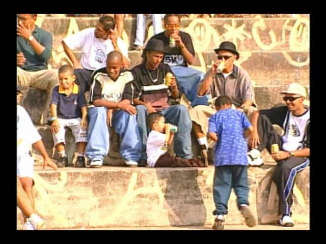 Foto do clipe 'Tik Tak', do Doctor Mc's, com os integrantes do grupo em meio jovens e alguns meninos sentados em uma arquibancada de cimento pichada, sob a luz do sol. As pessoas, em sua maioria negras, vestem roupas casuais e de estilo urbano, como calças largas, camisetas e bonés. A atmosfera é descontraída, e alguns deles seguram latas de bebida.