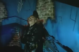 Douglas aparece no centro da imagem com expressão séria, sob uma iluminação dramática em um cenário escuro; ele veste uma jaqueta escura, aparentemente de couro. As paredes de tijolo ao fundo estão projetadas em tons fortes de azul, com sombras profundas e rastros de grafite ('VIDA' é visível no topo esquerdo). A iluminação intensa de baixo destaca seu rosto e cria um contraste marcante.