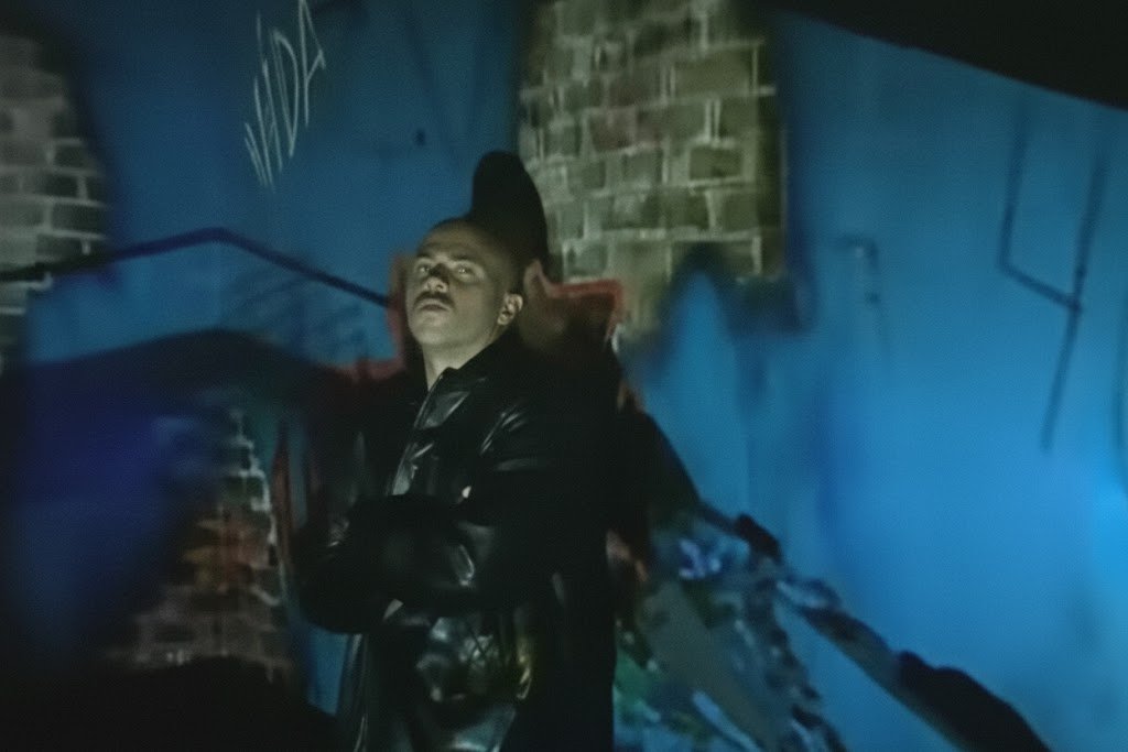 Douglas aparece no centro da imagem com expressão séria, sob uma iluminação dramática em um cenário escuro; ele veste uma jaqueta escura, aparentemente de couro. As paredes de tijolo ao fundo estão projetadas em tons fortes de azul, com sombras profundas e rastros de grafite ('VIDA' é visível no topo esquerdo). A iluminação intensa de baixo destaca seu rosto e cria um contraste marcante.