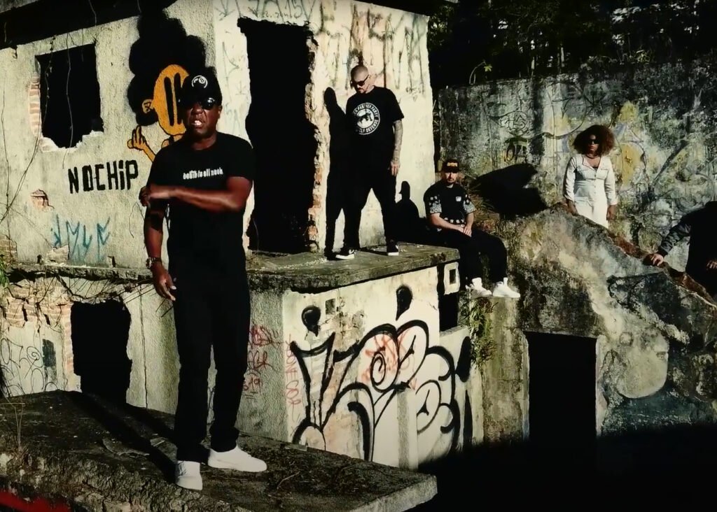 Membros do Realidade Cruel sob forte luz do sol em meio a paredes de concreto 'abandonadas', todas grafitada. A cena é do videoclipe 'A História Continua'. À frente, Markão II se destaca.
