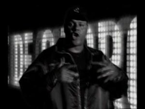 Uma captura de tela em preto e branco do videoclipe 'Círculo Vicioso', do Câmbio Negro. X está no centro, usando jaqueta e boné, gesticulando com as mãos enquanto canta. Ao fundo, um painel exibe letras grandes e luminosas de forma desfocada.