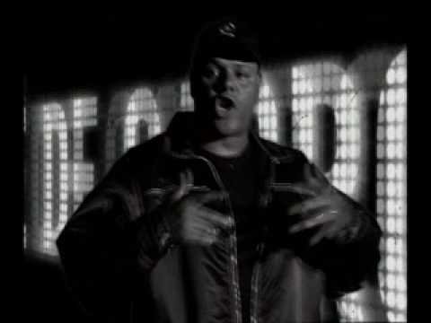 Uma captura de tela em preto e branco do videoclipe 'Círculo Vicioso', do Câmbio Negro. X está no centro, usando jaqueta e boné, gesticulando com as mãos enquanto canta. Ao fundo, um painel exibe letras grandes e luminosas de forma desfocada.