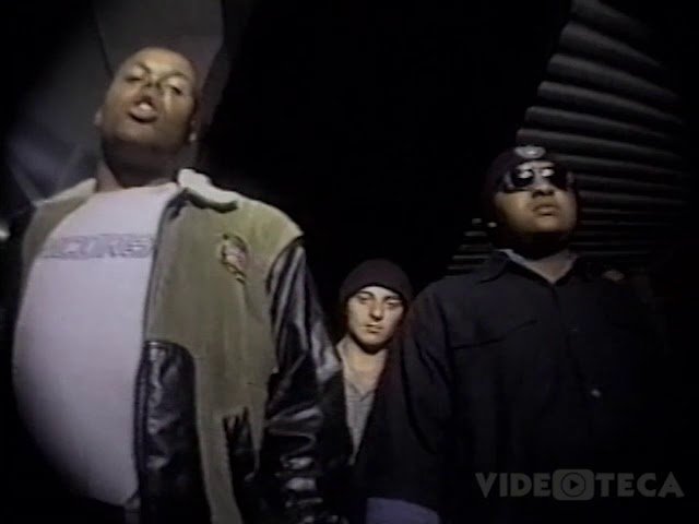 Uma cena de um videoclipe de baixa resolução, filmada de baixo para cima, mostrando três homens em um ambiente noturno. Em primeiro plano à esquerda, X (Câmbio Negro) canta com uma jaqueta de mangas de couro. À direita, DJ Jamaika usa óculos escuros e touca preta. Um terceiro homem, branco e de touca, está ao centro, um pouco atrás. A imagem tem uma estética de vídeo VHS.