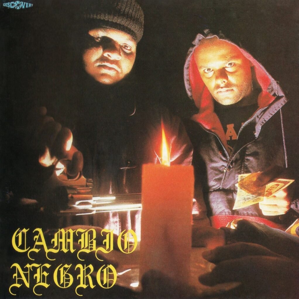 A capa do álbum 'Sub-Raça' do grupo Câmbio Negro. A foto, em um ambiente escuro, é centralizada em dois homens (os integrantes X e DJ Jamaika) iluminados por uma grande vela laranja acesa em primeiro plano, que está sobre uma mesa. DJ Jamaika, à esquerda, usa uma touca preta e X, à direita, usa um moletom escuro com capuz de forro vermelho, segurando o que parecem ser notas de dinheiro. Seus rostos são iluminados de baixo para cima pela chama, criando um ar sombrio e intenso. Na parte inferior esquerda, o nome 'CÂMBIO NEGRO' está escrito em uma fonte gótica amarela. No canto superior esquerdo, vê-se a palavra 'DISCOVERY'.