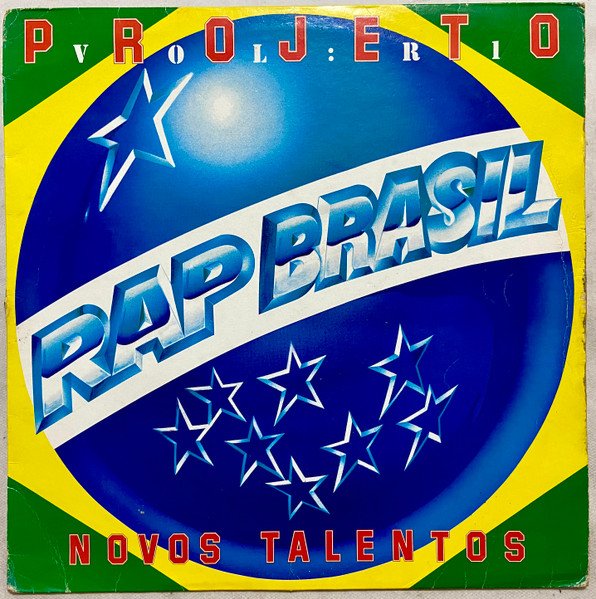 A capa do álbum 'Projeto Rap Brasil 1: Novos Talentos'. A arte é uma estilização da bandeira do Brasil. Ao centro, há um grande globo azul com estrelas brancas, cortado por uma faixa branca onde está escrito 'RAP BRASIL' em letras grandes, prateadas e com efeito 3D. Na parte superior, dentro da borda verde, lê-se 'PROJETO RAP BRASIL', e entre a palavra 'PROJETO' lê-se 'vol. 1'. Na parte inferior, em vermelho, está o subtítulo 'NOVOS TALENTOS'. A moldura da capa alterna entre o verde (topo e base) e o amarelo (laterais), mimetizando as cores da bandeira nacional.