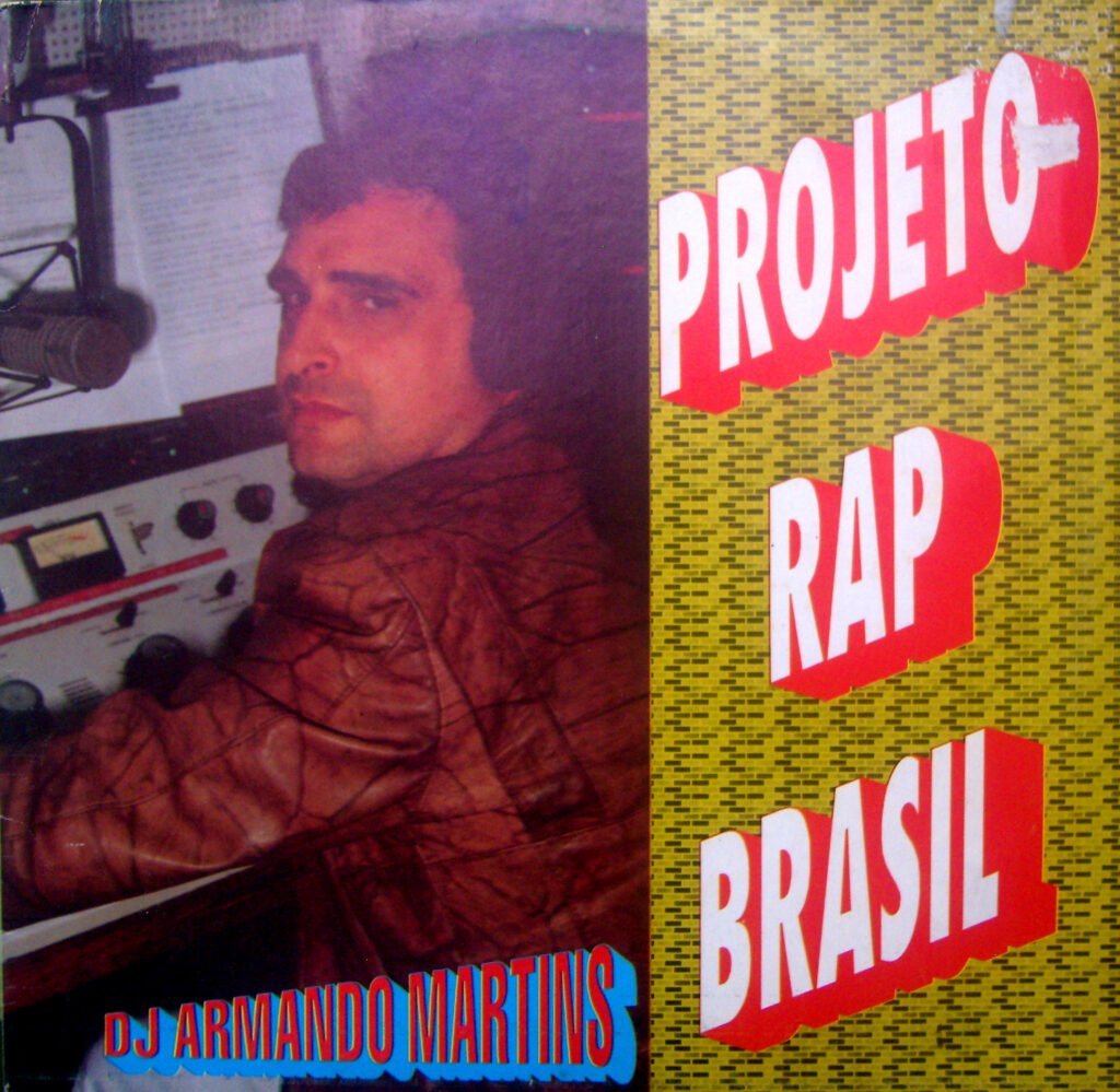 A capa do álbum 'Projeto Rap Brasil Vol. 2', também conhecido como 'Rap Mania', do DJ Armando Martins. A imagem é dividida: à esquerda, vê-se o DJ Armando Martins sentado em um estúdio de rádio, olhando por cima do ombro para a câmera. Ele veste uma jaqueta de couro marrom e está diante de uma mesa de som (mixer) e um microfone. Ao fundo, papéis com textos, possivelmente roteiros ou playlists, são visíveis. Na parte inferior esquerda, o nome 'DJ ARMANDO MARTINS' está escrito em letras azuis com contorno vermelho. À direita, sobre um fundo amarelo com um padrão gráfico repetitivo em preto, o título 'PROJETO RAP BRASIL' está disposto em três linhas, com letras grandes, vermelhas e sombreadas em branco, inclinadas para cima.