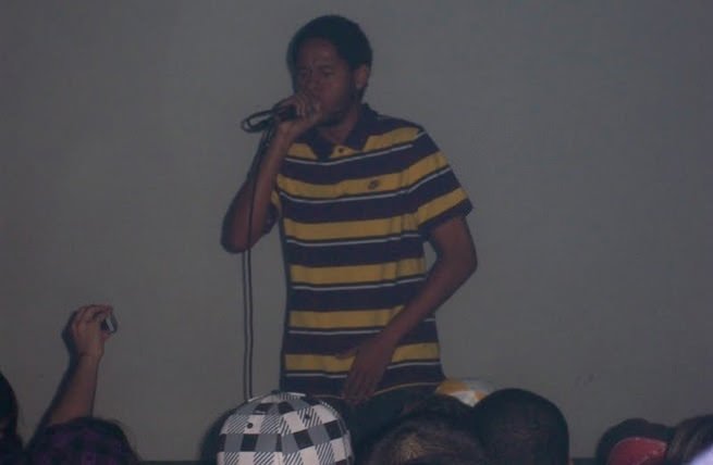 Fotografia de um show de rap em ambiente fechado e com pouca iluminação. Ao centro, o rapper Emicida, ainda jovem, aparece segurando um microfone próximo à boca com a mão direita, enquanto a mão esquerda está próxima à cintura. Ele veste uma camisa polo de listras horizontais largas nas cores amarelo e marrom escuro. O fundo é uma parede cinza lisa e mal iluminada. Na parte inferior da imagem, é possível ver o topo da cabeça de algumas pessoas da plateia e uma mão segurando uma câmera fotográfica pequena à esquerda, registrando o momento.