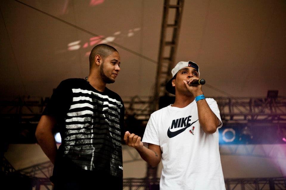 Emicida, à esquerda, olhando para Rashid, à direita, que segura o microfone em cima do palco. Emicida veste uma camisa preta enquanto Rashid veste uma branca com o logo da Nike em destaque.