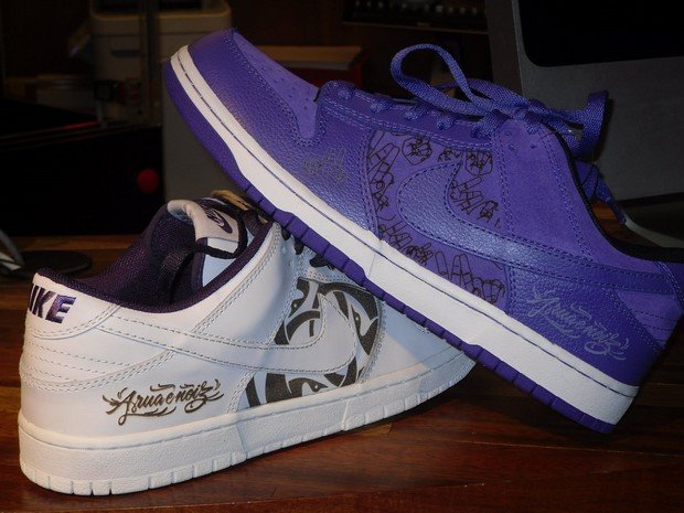 Par de tênis Nike com o pé esquerdo, em roxo, por cima do pé direito, branco. Inscrições de "A rua é nóiz" personalizadas aparecem nos pés.
