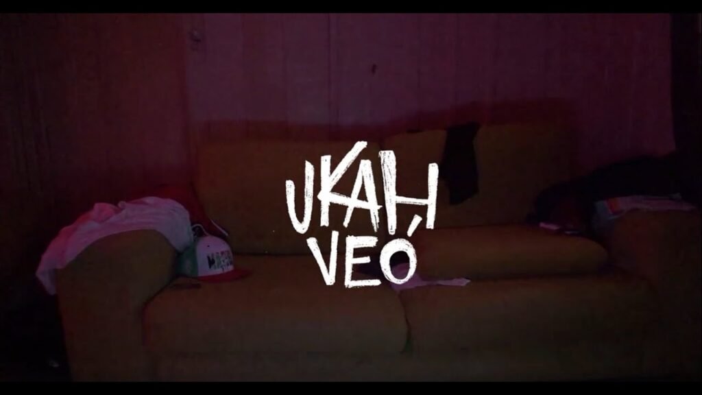 Uma captura de ecrã de um videoclipe ambientado num interior escurecido com iluminação avermelhada e sombria. No centro da imagem, um sofá de cor ocre ou mostarda está posicionado contra uma parede de madeira. Sobre o sofá, à esquerda, vê-se um boné de aba reta verde e branco e algumas peças de roupa desalinhadas. Sobrepostas ao centro do sofá, as palavras 'UKAH,' e 'VEO' aparecem escritas em letras brancas estilizadas com uma textura que lembra giz ou tinta desgastada. A atmosfera geral da cena é urbana, caseira e intimista.