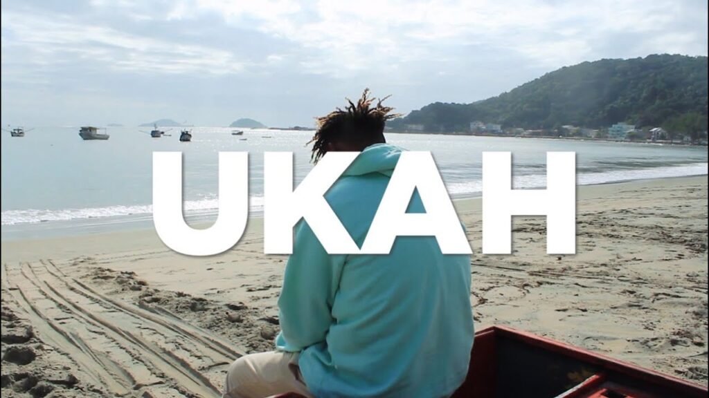 Uma captura de ecrã de um videoclipe gravado numa praia durante um dia claro. No centro, o rapper Ukah aparece de costas, sentado na areia ou numa pequena embarcação, vestindo um moletom com capuz de cor azul-bebê e calças beges. À sua frente, o mar calmo exibe vários barcos de pesca ancorados perto da costa. Ao fundo, à direita, destaca-se uma encosta verdejante com algumas casas na base. Sobreposta ao centro da imagem, a palavra 'UKAH' aparece escrita em letras garrafais brancas e sólidas.