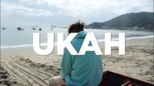 Uma captura de ecrã de um videoclipe gravado numa praia durante um dia claro. No centro, o rapper Ukah aparece de costas, sentado na areia ou numa pequena embarcação, vestindo um moletom com capuz de cor azul-bebê e calças beges. À sua frente, o mar calmo exibe vários barcos de pesca ancorados perto da costa. Ao fundo, à direita, destaca-se uma encosta verdejante com algumas casas na base. Sobreposta ao centro da imagem, a palavra 'UKAH' aparece escrita em letras garrafais brancas e sólidas.