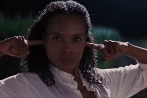 Uma mulher negra e cabelos escuros cacheados, na altura dos ombros, encara a câmera com uma expressão séria e decidida. Ela veste uma camisa branca de botões e está com as mãos erguidas, usando os dedos indicadores para tapar os ouvidos. O fundo está escurecido e levemente fora de foco, sugerindo um ambiente noturno ao ar livre com folhagens. A iluminação destaca o rosto da mulher e a textura de seu cabelo.