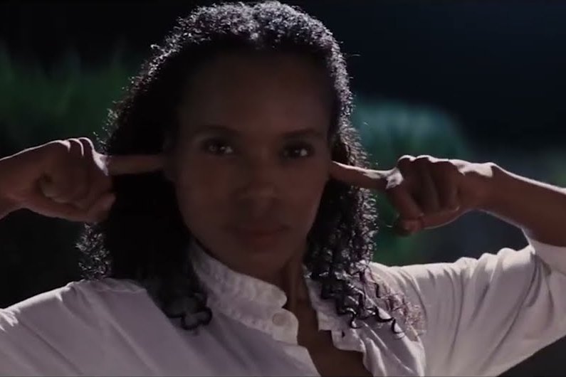 Uma mulher negra e cabelos escuros cacheados, na altura dos ombros, encara a câmera com uma expressão séria e decidida. Ela veste uma camisa branca de botões e está com as mãos erguidas, usando os dedos indicadores para tapar os ouvidos. O fundo está escurecido e levemente fora de foco, sugerindo um ambiente noturno ao ar livre com folhagens. A iluminação destaca o rosto da mulher e a textura de seu cabelo.