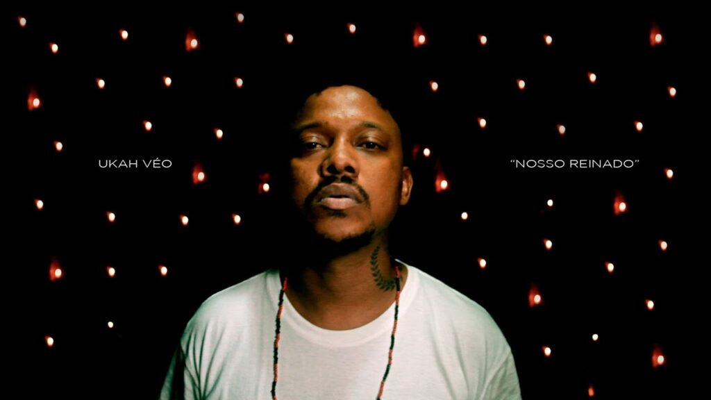 Uma captura de ecrã de um videoclipe com estética noturna e intimista. No centro, o rapper Ukah aparece num grande plano frontal, olhando seriamente para a câmara. Ele veste uma t-shirt branca simples, usa um colar de contas vermelhas e pretas e tem uma tatuagem de ramo de louros no lado direito do pescoço. O fundo é completamente preto, decorado com vários pontos de luz desfocados (efeito bokeh) que lembram pequenas velas ou luzes de Natal. À esquerda do artista, lê-se 'UKAH VÉO' e, à direita, o título 'NOSSO REINADO' entre aspas, ambos escritos em letras brancas pequenas e finas.