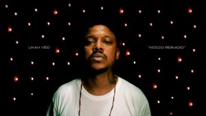 Uma captura de ecrã de um videoclipe com estética noturna e intimista. No centro, o rapper Ukah aparece num grande plano frontal, olhando seriamente para a câmara. Ele veste uma t-shirt branca simples, usa um colar de contas vermelhas e pretas e tem uma tatuagem de ramo de louros no lado direito do pescoço. O fundo é completamente preto, decorado com vários pontos de luz desfocados (efeito bokeh) que lembram pequenas velas ou luzes de Natal. À esquerda do artista, lê-se 'UKAH VÉO' e, à direita, o título 'NOSSO REINADO' entre aspas, ambos escritos em letras brancas pequenas e finas.