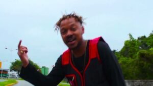 Uma captura de ecrã de um videoclipe ao ar livre durante um dia nublado. No centro da imagem, o rapper Ukah aparece em plano médio, olhando e apontando o dedo indicador da mão direita para cima. Ele tem o cabelo curto com dreadlocks loiros e veste um colete preto com detalhes em vermelho vibrante sobre uma t-shirt preta de manga comprida. Ao fundo, à esquerda, vê-se uma avenida com postes de iluminação e, à direita, uma encosta com vegetação densa e árvores verdes. A imagem tem um aspeto ligeiramente desfocado, capturando o movimento do artista.