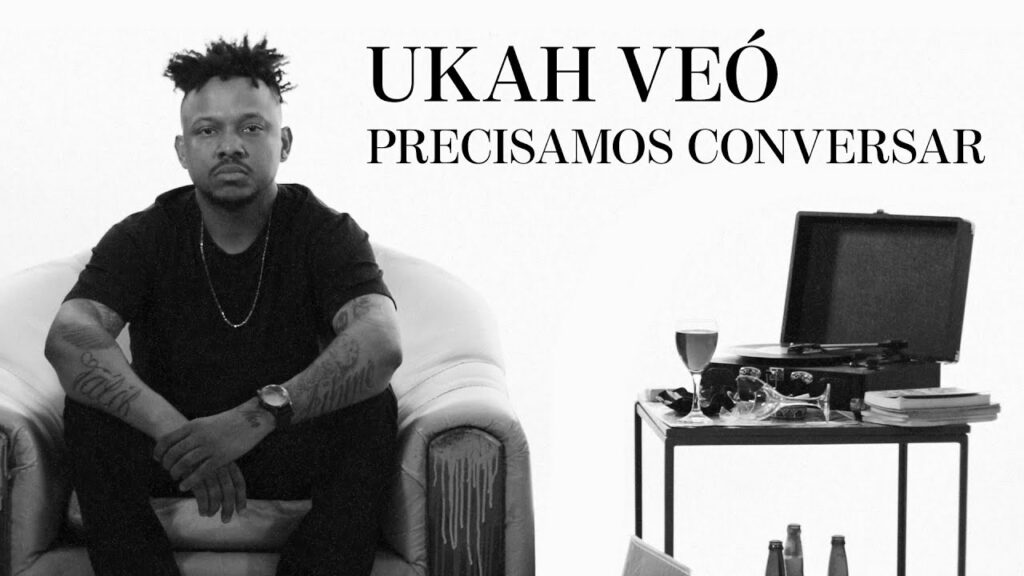 Uma imagem em preto e branco mostra o artista Ukah Veó sentado em uma poltrona de couro clara, posicionado à esquerda. Ele é um homem negro com cabelo estilo 'dreadlocks' curtos para cima, usa uma camiseta preta, colar de corrente fina, relógio no pulso esquerdo e tem tatuagens visíveis nos antebraços. À direita, há uma mesa lateral com um toca-discos aberto, uma taça com bebida, um copo quebrado e alguns livros empilhados. No canto superior direito, o texto em letras maiúsculas diz: 'UKAH VEÓ' e, logo abaixo, 'PRECISAMOS CONVERSAR'. O fundo é inteiramente branco e minimalista.