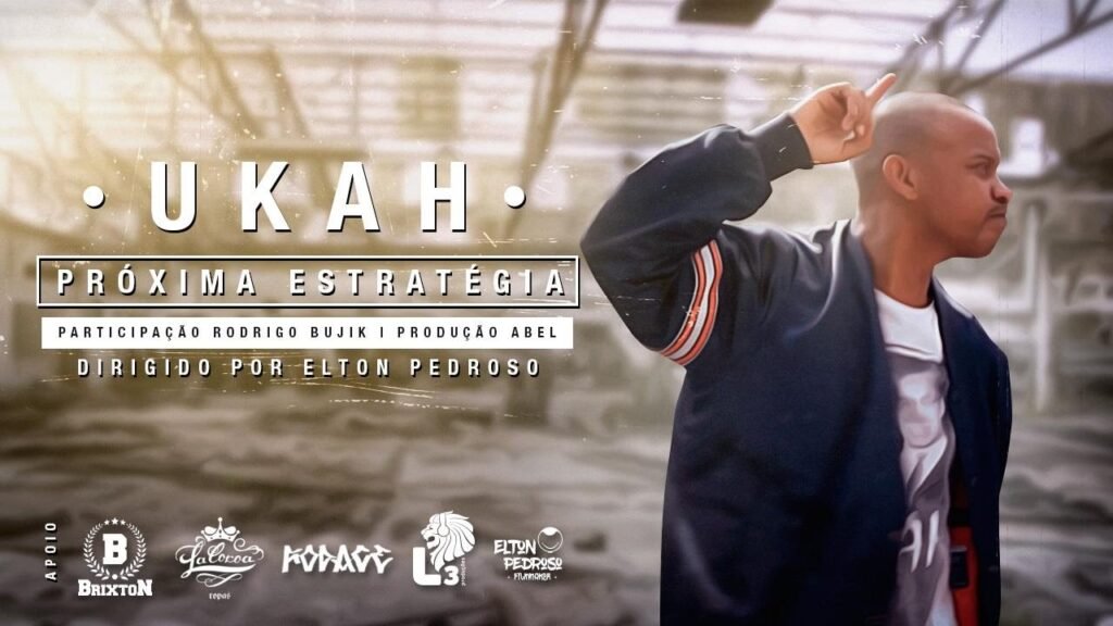 Uma captura de ecrã de um videoclipe com uma estética envelhecida e desbotada. À direita, o rapper Ukah aparece de perfil, com a cabeça rapada, vestindo um casaco desportivo azul escuro com riscas cor de laranja e brancas na manga. Ele tem o braço direito levantado, apontando o dedo indicador para cima. À esquerda, sobre um fundo desfocado que sugere um ambiente industrial ou um armazém, aparecem os créditos do vídeo em letras brancas: o nome 'UKAH' no topo, seguido pelo título 'PRÓXIMA ESTRATÉGIA' dentro de um retângulo, e as informações de participação de Rodrigo Bujik, produção de Abel e direção de Elton Pedroso. Na parte inferior, alinham-se vários logótipos de apoios, incluindo 'BRIXTON', 'La Coroa' e 'Elton Pedroso Filmmaker'.