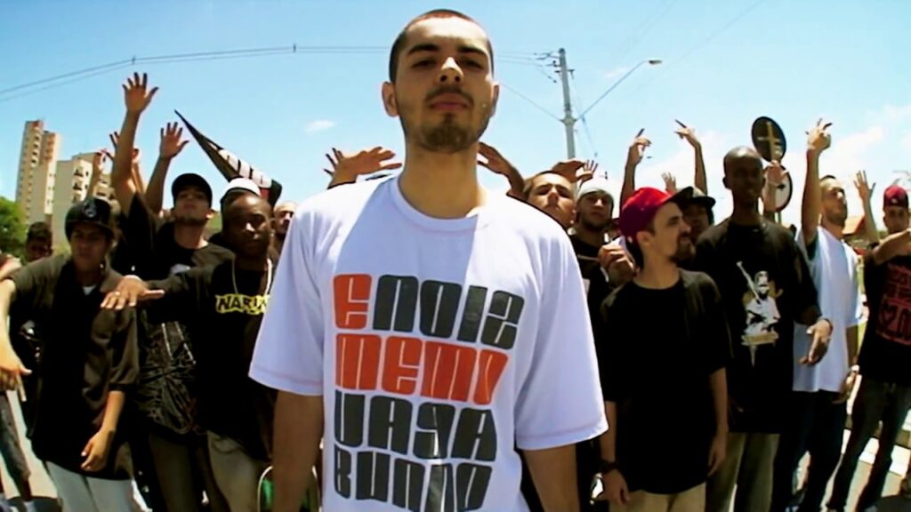 Fotografia com efeito de lente grande angular (fisheye) tirada ao ar livre em um dia ensolarado. Em primeiro plano, no centro da imagem, está o rapper A.X.L., um jovem de pele clara, cabelo escuro bem curto e cavanhaque, olhando diretamente para a câmera com uma expressão séria. Ele veste uma camiseta branca folgada com uma estampa em letras de forma grandes e quadradas nas cores laranja e preto, formando a frase 'e noiz memo vagabundo'. Atrás dele, há uma multidão de jovens homens, muitos erguendo os braços e fazendo gestos com as mãos típicos da cultura hip-hop. O cenário urbano ao fundo exibe postes de energia elétrica, placas de trânsito, algumas árvores e prédios distantes sob um céu azul claro.