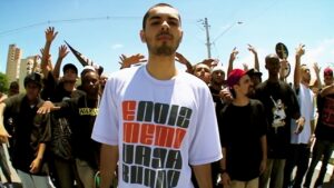 Fotografia com efeito de lente grande angular (fisheye) tirada ao ar livre em um dia ensolarado. Em primeiro plano, no centro da imagem, está o rapper A.X.L., um jovem de pele clara, cabelo escuro bem curto e cavanhaque, olhando diretamente para a câmera com uma expressão séria. Ele veste uma camiseta branca folgada com uma estampa em letras de forma grandes e quadradas nas cores laranja e preto, formando a frase 'e noiz memo vagabundo'. Atrás dele, há uma multidão de jovens homens, muitos erguendo os braços e fazendo gestos com as mãos típicos da cultura hip-hop. O cenário urbano ao fundo exibe postes de energia elétrica, placas de trânsito, algumas árvores e prédios distantes sob um céu azul claro.