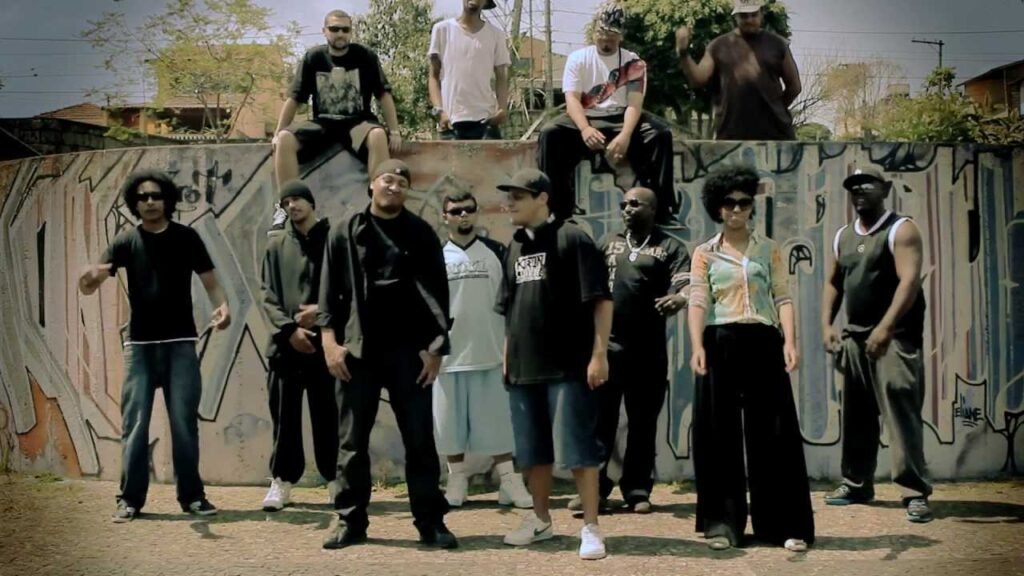 A cena do clipe da música 'Perigo', produzida pelo DJ Nato PK, registra uma reunião de peso que simboliza a força do coletivo e do selo Pau-de-Dá-em-Doido. Além do DJ, o quadro reúne os MCs André Sagat, Max Musicamente, Preto-R, Enézimo, Eri Q.i e Stefanie, que também emprestam suas vozes e líricas à faixa. Posicionados em formação de grupo à frente de um extenso muro com 'graffitis' e 'tags', os artistas reforçam a ocupação urbana e a estética clássica do hip hop de rua.